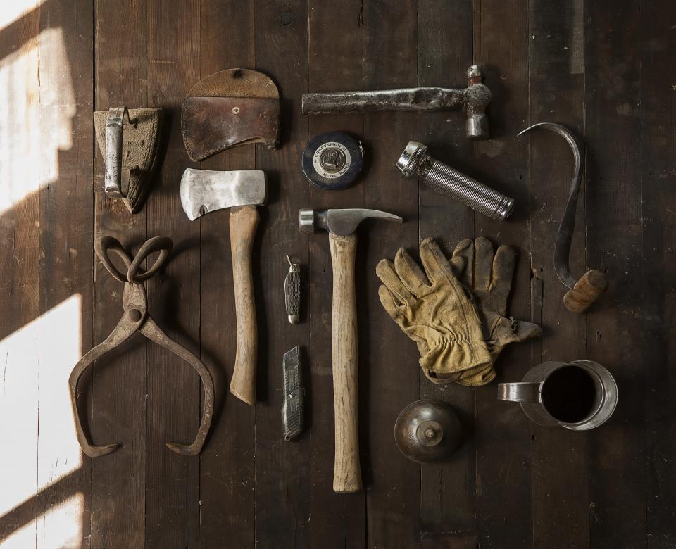 An overhead flat lay of well-used hand tools — a hatchet, hammers, tin snips, pocket knives and a flashlight — arranged in rows