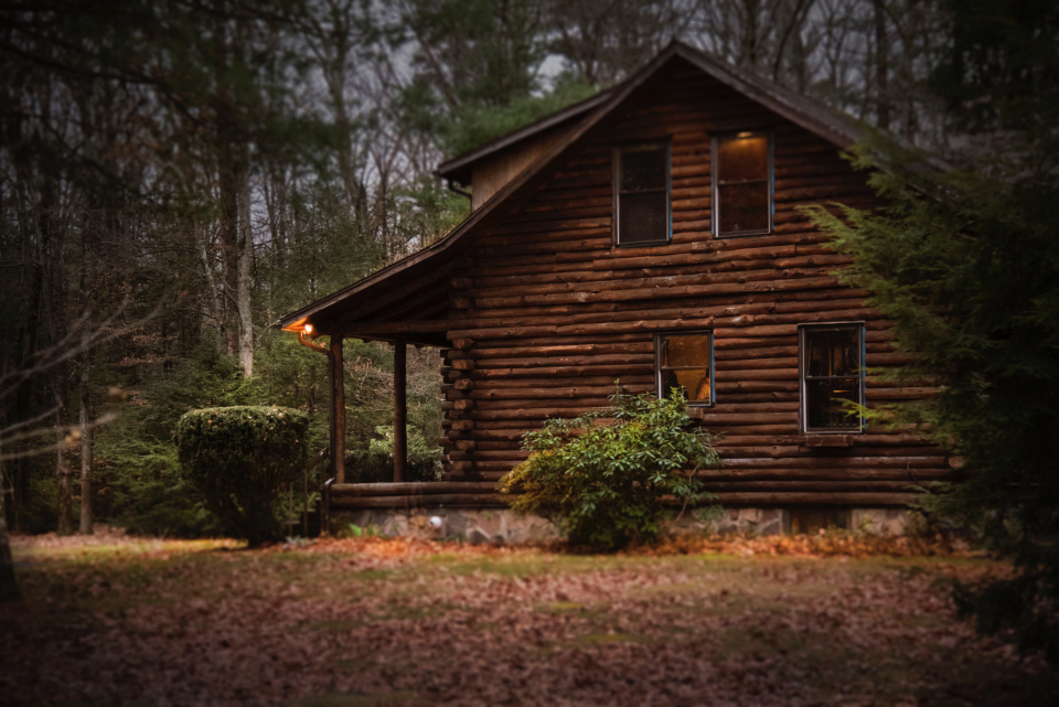 A log cabin among bare winter trees at dusk, warm light glowing from an eave lamp and the upstairs windows
